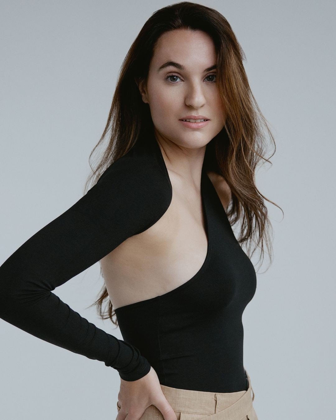 Woman with long brown hair wears a black, one-sleeve, asymmetrical top and beige pants, standing against a plain light gray background.
